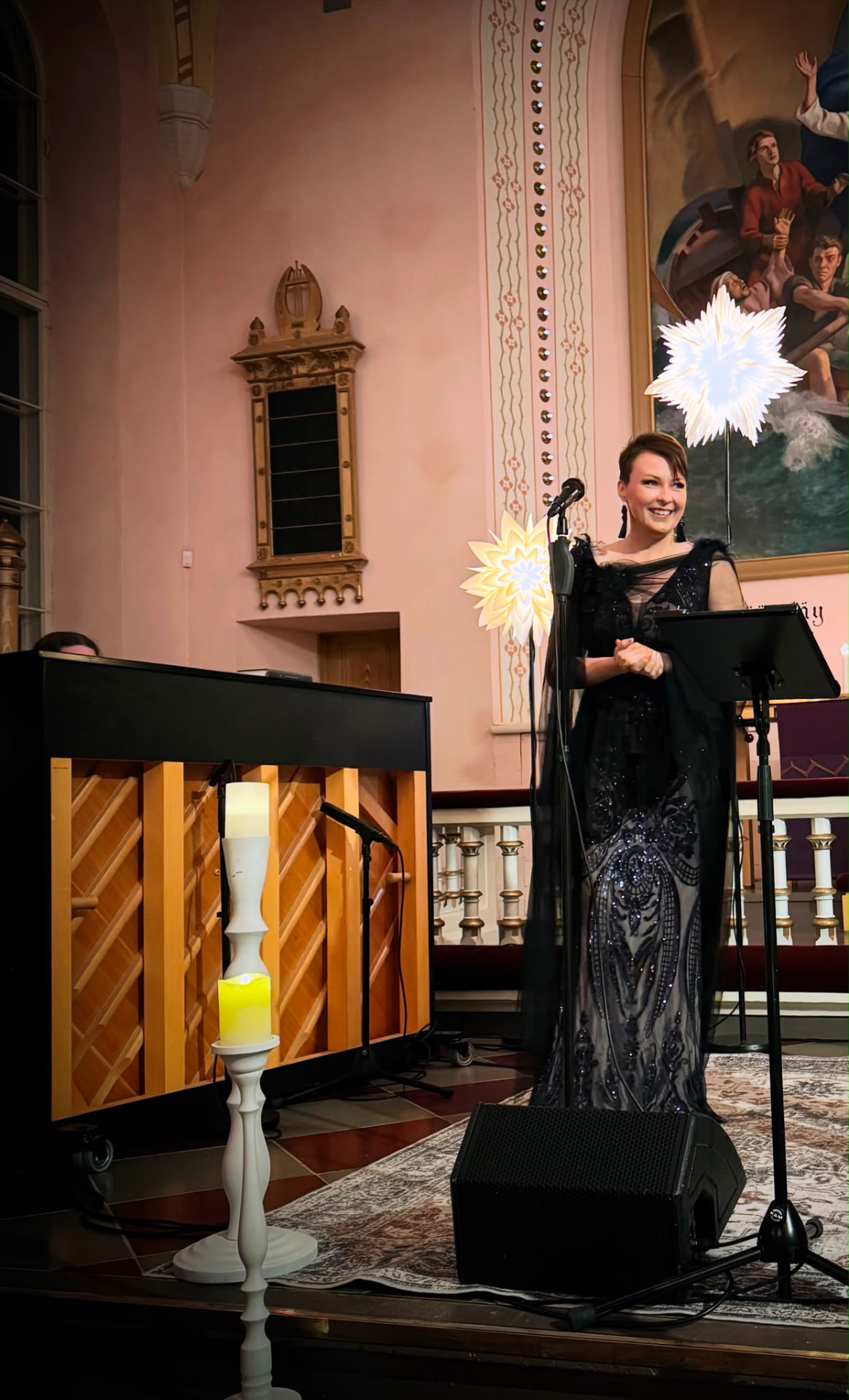 Maikki laulaa mustassa juhlapuvussa kirkkokonsertissa kynttilöiden ja valotähtien loisteessa